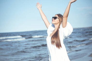 Mutlu sarışın kadın okyanus sahilinde beyaz bir elbise ve güneş gözlüğü ile el kaldırıyor..