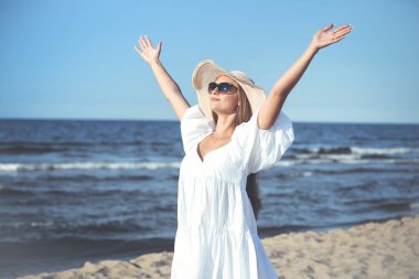 Mutlu sarışın kadın okyanus sahilinde beyaz elbiseli, güneş gözlüklü ve şapkalı el kaldırıyor..