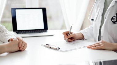Doktor ve hasta klinikteki beyaz masada yan yana oturuyorlar. Kadın hekim bir kayıt formunu doldururken dinliyor. Tıp konsepti.