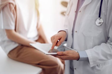 Doktor ve hasta klinikte. Dost canlısı bir doktor genç bir kadının yanında tablet bilgisayar kullanıyor. Tıp konsepti.