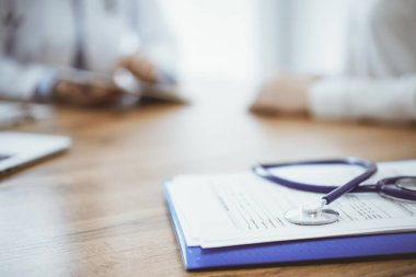 Stetoskop ve ilaç geçmişi kayıtları ahşap masada yatarken doktor ve hasta arka planda oturup birbirleriyle konuşuyorlar. İlaç..
