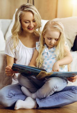 Mutlu bir aile. Sarışın genç anne güneşli odada ahşap zeminde otururken sevimli kızına kitap okuyor. Annelik kavramı.