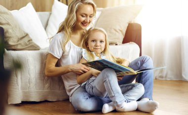 Mutlu bir aile. Sarışın genç anne güneşli odada ahşap zeminde otururken sevimli kızına kitap okuyor. Annelik kavramı.