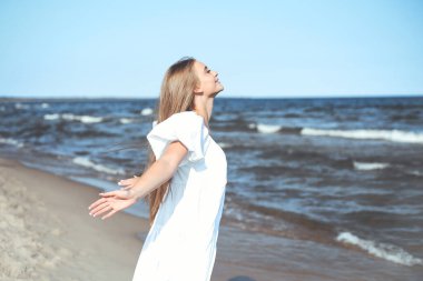 Mutlu gülümseyen güzel kadın okyanus sahilinde beyaz bir yaz elbisesi içinde, kolları açık..