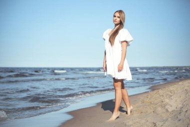 Mutlu gülümseyen güzel kadın beyaz bir yaz elbisesiyle okyanus sahilinde yürüyor..