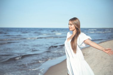 Okyanus sahilinde beyaz bir yaz elbisesi içinde kollarını açmış mutlu, güzel bir kadın..