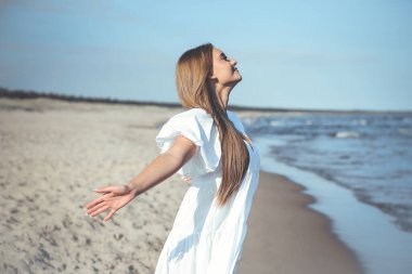 Mutlu gülümseyen güzel kadın okyanus sahilinde beyaz bir yaz elbisesi içinde, kolları açık..