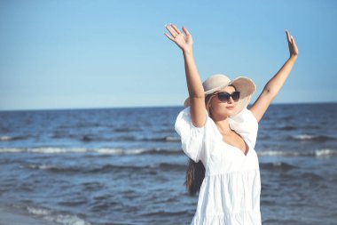 Mutlu sarışın kadın okyanus sahilinde beyaz bir elbise ve güneş gözlüğü ile el kaldırıyor..