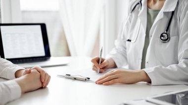 Doktor ve hasta klinikteki beyaz masada yan yana oturuyorlar. Kadın hekim bir kayıt formunu doldururken dinliyor. Tıp konsepti.