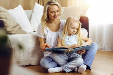 Mutlu bir aile. Sarışın genç anne güneşli odada ahşap zeminde otururken sevimli kızına kitap okuyor. Annelik kavramı.