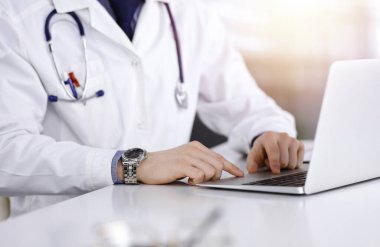 Bilinmeyen bir erkek doktor karanlık bir klinikte dizüstü bilgisayarla oturuyor ve çalışıyor. Arka planda ışık parıltısı var..