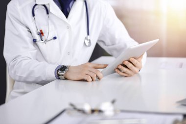 Bilinmeyen erkek doktor, tablet bilgisayarla oturuyor ve çalışıyor. Karanlık bir klinikte, arka planda ışık parıltısı, ellerin yakın çekimi..