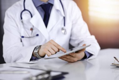 Bilinmeyen erkek doktor, tablet bilgisayarla oturuyor ve çalışıyor. Karanlık bir klinikte, arka planda ışık parıltısı, ellerin yakın çekimi..