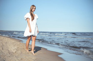 Mutlu gülümseyen güzel kadın beyaz bir yaz elbisesiyle okyanus sahilinde yürüyor..