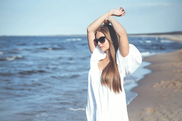 Happy blonde woman is on the ocean beach in a white dress and sunglasses, raising hands.