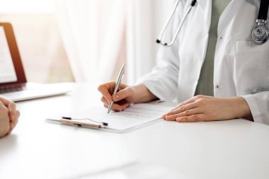 Doktor ve hasta klinikteki beyaz masada yan yana oturuyorlar. Kadın hekim bir kayıt formunu doldururken dinliyor. Tıp konsepti.