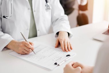 Doktor ve hasta klinikteki beyaz masada yan yana oturuyorlar. Kadın hekim bir kayıt formunu doldururken dinliyor. Tıp konsepti.