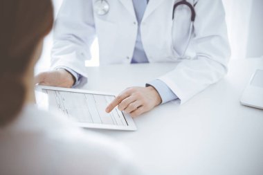 Doktor ve hasta klinikteki masada karşılıklı oturuyorlar. Kadın doktorların elleri tablet bilgisayarın dokunmatik dokunmatik pedini işaret ediyor, kapat. Tıp konsepti.