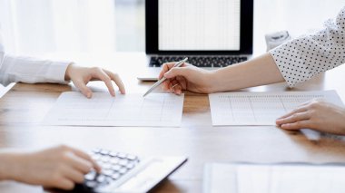 Muhasebeci kadın, ofisteki ahşap masada meslektaşıyla birlikte vergi sayarken hesap makinesi ve dizüstü bilgisayar kullanıyor. İş denetimi ve finans alanında takım çalışması.
