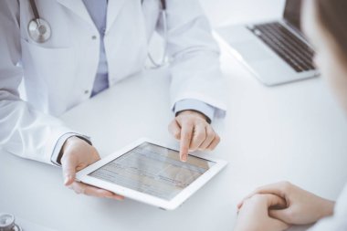 Doktor ve hasta klinikteki masada karşılıklı oturuyorlar. Kadın doktorların elleri tablet bilgisayarın dokunmatik dokunmatik pedini işaret ediyor, kapat. Tıp konsepti.