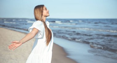 Okyanus sahilinde beyaz bir yaz elbisesi içinde kollarını açmış mutlu, güzel bir kadın..