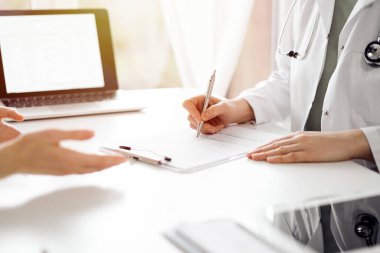 Doktor ve hasta klinikteki beyaz masada oturup bir şeyler tartışıyorlar. Kadın hekim bir kayıt formunu doldururken dinliyor. Tıp konsepti.
