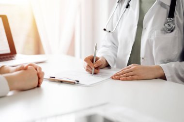 Doktor ve hasta klinikteki beyaz masada yan yana oturuyorlar. Kadın hekim bir kayıt formunu doldururken dinliyor. Tıp konsepti.
