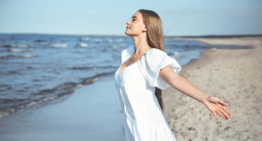 Mutlu gülümseyen güzel kadın okyanus sahilinde beyaz bir yaz elbisesi içinde, kolları açık..