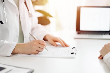 Doktor ve hasta klinikteki beyaz masada yan yana oturuyorlar. Kadın hekim bir kayıt formunu doldururken dinliyor. Tıp konsepti.