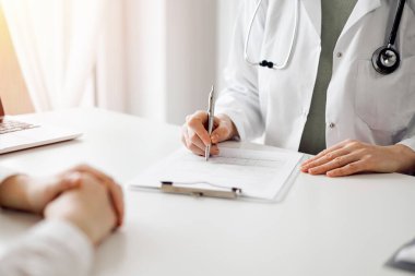 Doktor ve hasta klinikteki beyaz masada yan yana oturuyorlar. Kadın hekim bir kayıt formunu doldururken dinliyor. Tıp konsepti.