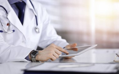 Bilinmeyen erkek doktor, tablet bilgisayarla oturuyor ve çalışıyor. Karanlık bir klinikte, arka planda ışık parıltısı, ellerin yakın çekimi..