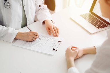 Doktor ve hasta klinikteki beyaz masada yan yana oturuyorlar. Kadın hekim bir kayıt formunu doldururken dinliyor. Tıp konsepti.