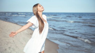 Okyanus sahilinde beyaz bir yaz elbisesi içinde kollarını açmış mutlu, güzel bir kadın..