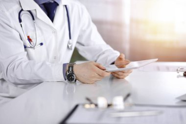 Bilinmeyen erkek doktor, tablet bilgisayarla oturuyor ve çalışıyor. Karanlık bir klinikte, arka planda ışık parıltısı, ellerin yakın çekimi..