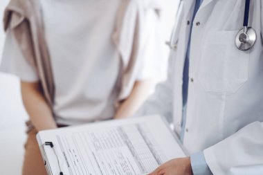 Doktor ve hasta klinikte. Bir yandan dostane bir doktor genç bir kadını muayene ederken diğer yandan tıbbi kayıtlarla dolu bir pano tutuyor. Tıp konsepti.