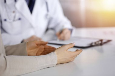 Unknown male doctor and patient woman discussing something while sittingin a darkened clinic, glare of light on the background. Close-up of hands. clipart