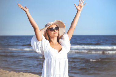 Mutlu sarışın kadın okyanus sahilinde beyaz elbiseli, güneş gözlüklü ve şapkalı el kaldırıyor..