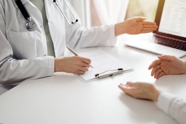 Doktor ve hasta klinikteki beyaz masada oturup bir şeyler tartışıyorlar. Kadın doktor dizüstü bilgisayarın ekranına işaret ediyor. Tıp konsepti.