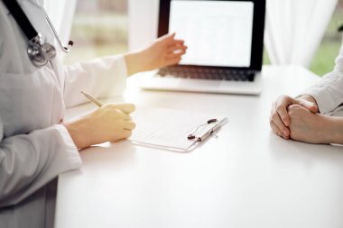 Doktor ve hasta klinikteki beyaz masada oturup bir şeyler tartışıyorlar. Kadın doktor dizüstü bilgisayarın ekranına işaret ediyor. Tıp konsepti.