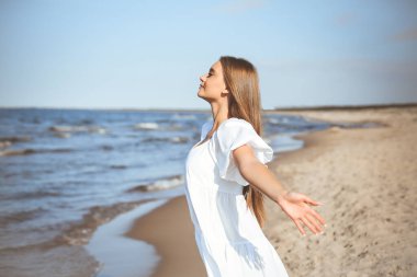 Mutlu gülümseyen güzel kadın okyanus sahilinde beyaz bir yaz elbisesi içinde, kolları açık..