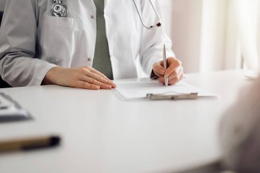 Doktor ve hasta klinikteki beyaz masada yan yana oturuyorlar. Kadın hekim bir kayıt formunu doldururken dinliyor. Tıp konsepti.