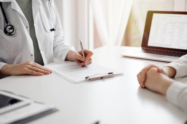 Doktor ve hasta klinikteki beyaz masada yan yana oturuyorlar. Kadın hekim bir kayıt formunu doldururken dinliyor. Tıp konsepti.