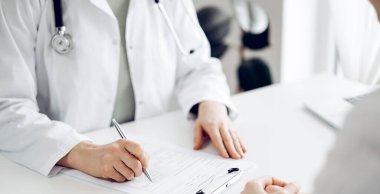 Doktor ve hasta klinikteki beyaz masada yan yana oturuyorlar. Kadın hekim bir kayıt formunu doldururken dinliyor. Tıp konsepti.