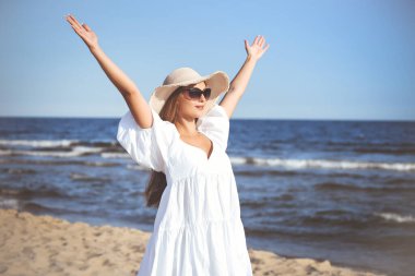 Mutlu sarışın kadın okyanus sahilinde beyaz elbiseli, güneş gözlüklü ve şapkalı el kaldırıyor..