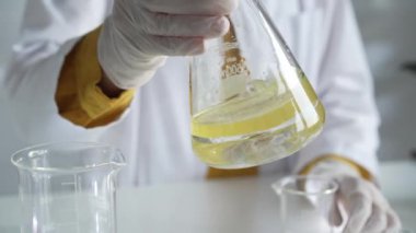 Beyaz koruyucu eldiven giyen bilinmeyen bilim adamı, bir laboratuarda, bir Erlenmeyer şişesinin içinde sarı bir kimyasal çözeltiyi dikkatlice sallıyor. Bilim ve tıp.
