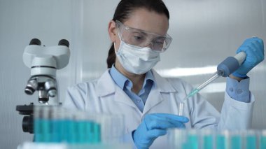 Laboratuvar önlüğü, maske, mavi eldiven ve güvenlik gözlüğü takan bilim kadını araştırmacı kadın, test tüpüne sıvı atmak için bir mikro pipet kullanıyor, portre görüntüsü. Tıp ve bilim konsepti.