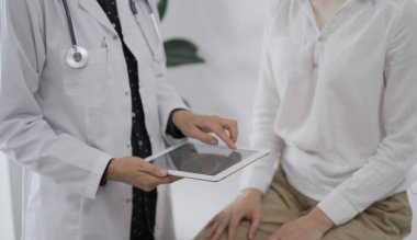 Doktor ve bir hasta bir şey tartışıyorlar. Kadın hekim, klinikteki muayene sırasında genç bir kadının yanında tıbbi notlarla tablet bilgisayar ekranına işaret ediyor..