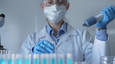 Laboratuvar önlüğü, maske, mavi eldiven ve güvenlik gözlüğü takan bilim adamı kadın test tüplerine sıvı atmak için mikropipet kullanıyor. Tıp ve Bilim.