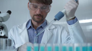 Laboratuvar önlüğü ve güvenlik gözlüğü takan bilim adamı deney tüpüne sıvı atmak için mikropipet kullanıyor. Portre görüntüsü. Tıp ve Bilim.