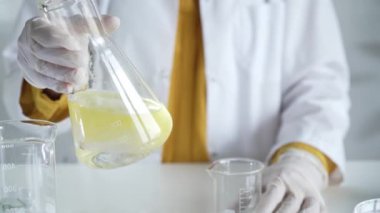 Beyaz koruyucu eldiven giyen bilinmeyen bilim adamı, bir laboratuarda, bir Erlenmeyer şişesinin içinde sarı bir kimyasal çözeltiyi dikkatlice sallıyor. Bilim ve tıp.
